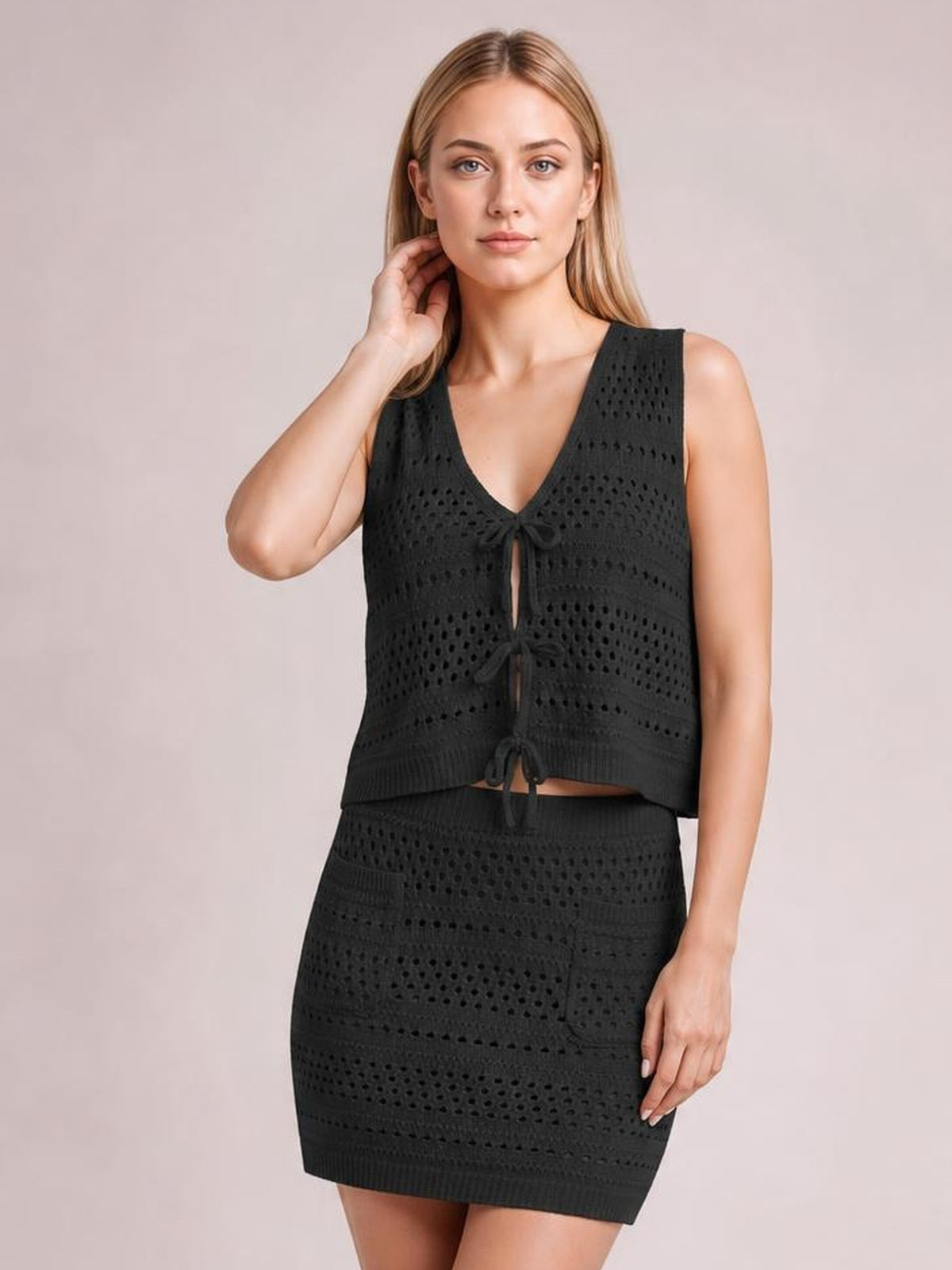 Model poses with one hand near her neck in a black openwork V-neck knit top and matching pocketed fitted skirt with tied front details.