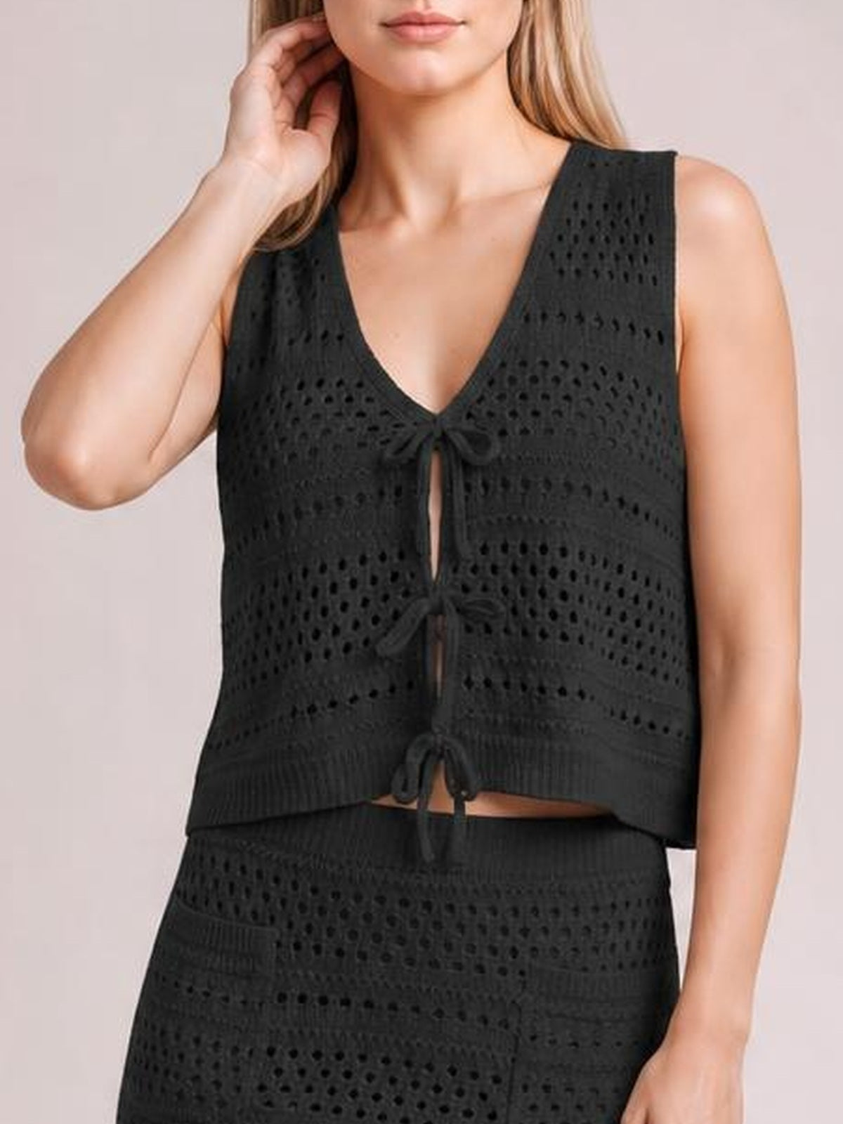 Model wears a black openwork knit cover-up set with a sleeveless V-neck top, front ties, cropped silhouette, and matching textured skirt.