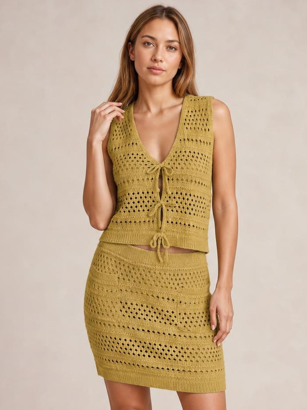 Model poses in a mustard openwork knit cover-up set with a sleeveless V-neck tied top and matching fitted mini skirt with pocket detail.