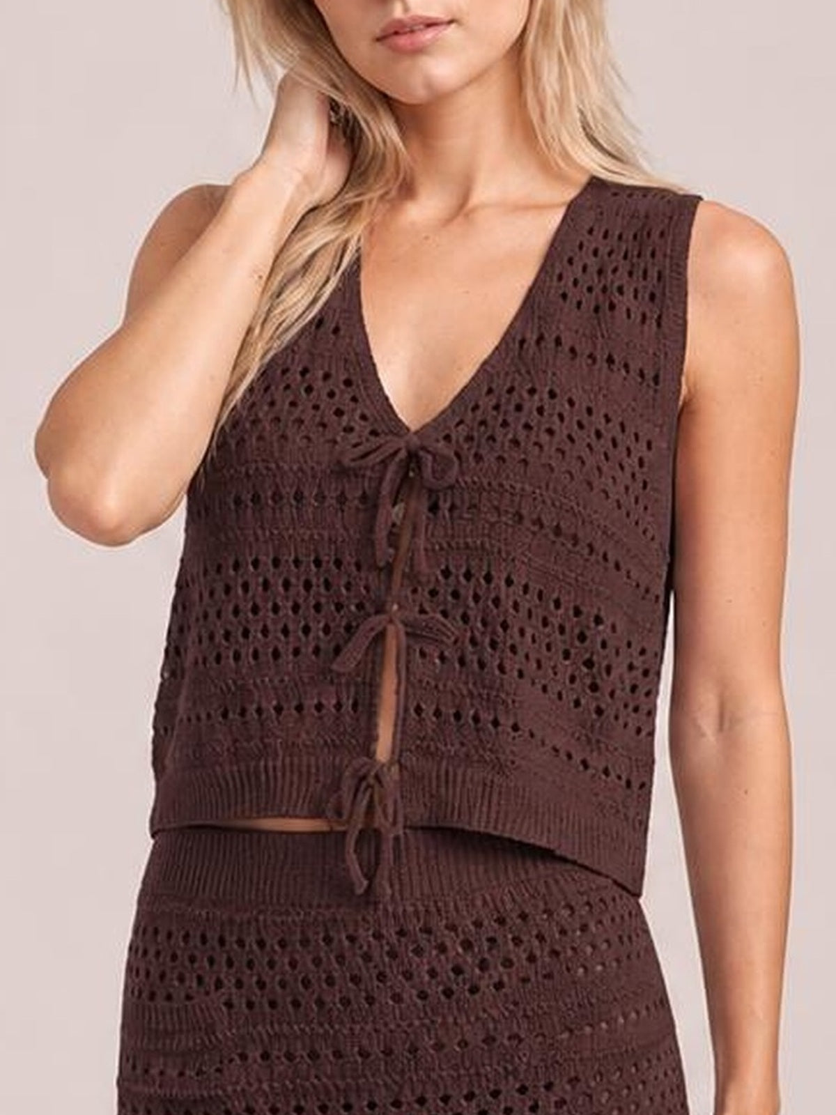Model poses with one hand in her hair wearing a dark brown openwork V-neck knit top with tied front, ribbed bands, and a matching skirt.