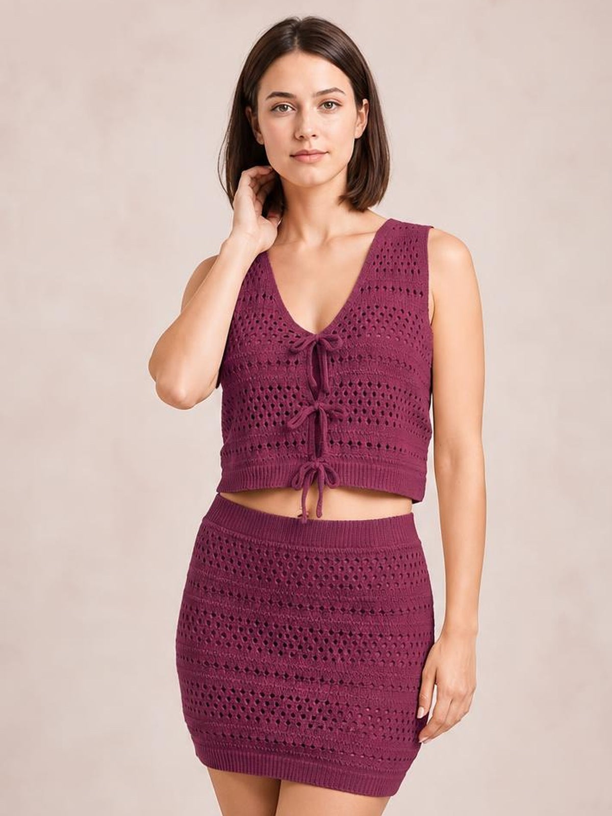 Model wearing a burgundy openwork knit cover-up set with a tied V-neck crop top and fitted mini skirt, posing with one hand near her neck.