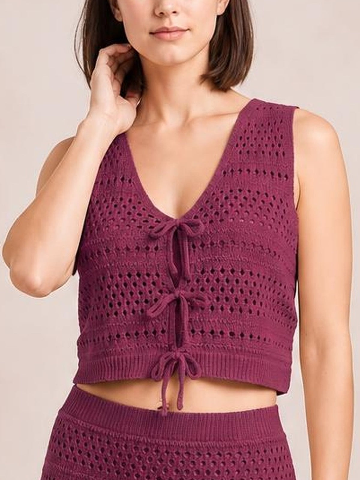 A model touches her hair while wearing a burgundy openwork knit cover-up set with a tied V-neck cropped top and matching ribbed skirt.