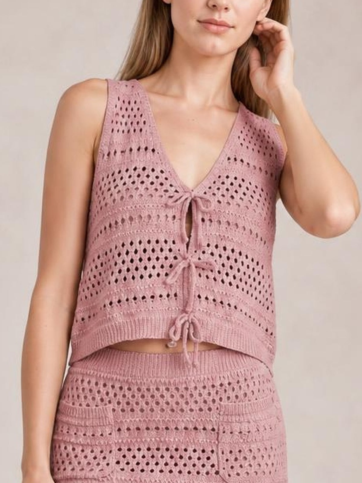 Model wearing a dusty pink openwork knit cover-up set with a tied V-neck top and matching high-waisted skirt, posing with one hand by her hair.
