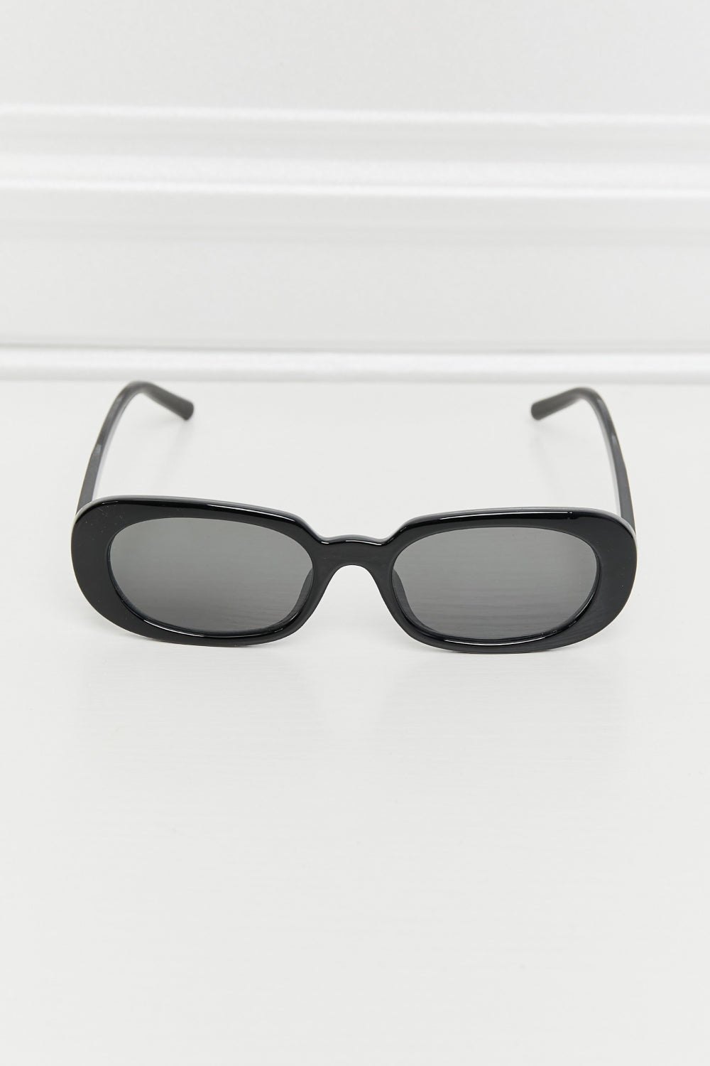 Black oval full-rim sunglasses feature softly curved dark lenses, a polished frame, and slim temples shown in a front-facing display.