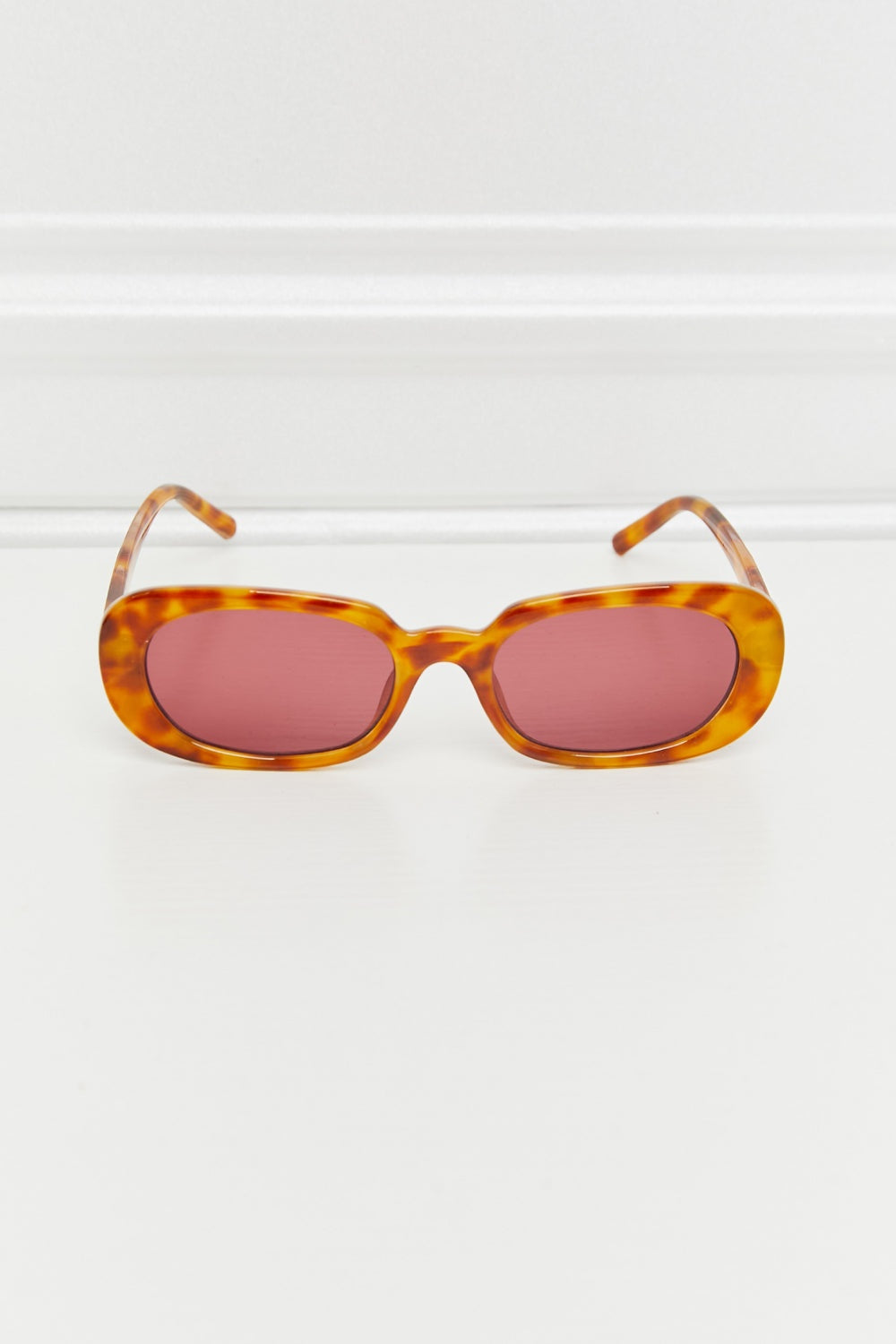 Tortoiseshell oval full-rim sunglasses feature softly curved pink lenses, a polished amber-brown frame, and slim temples folded open for a vintage-inspired silhouette.
