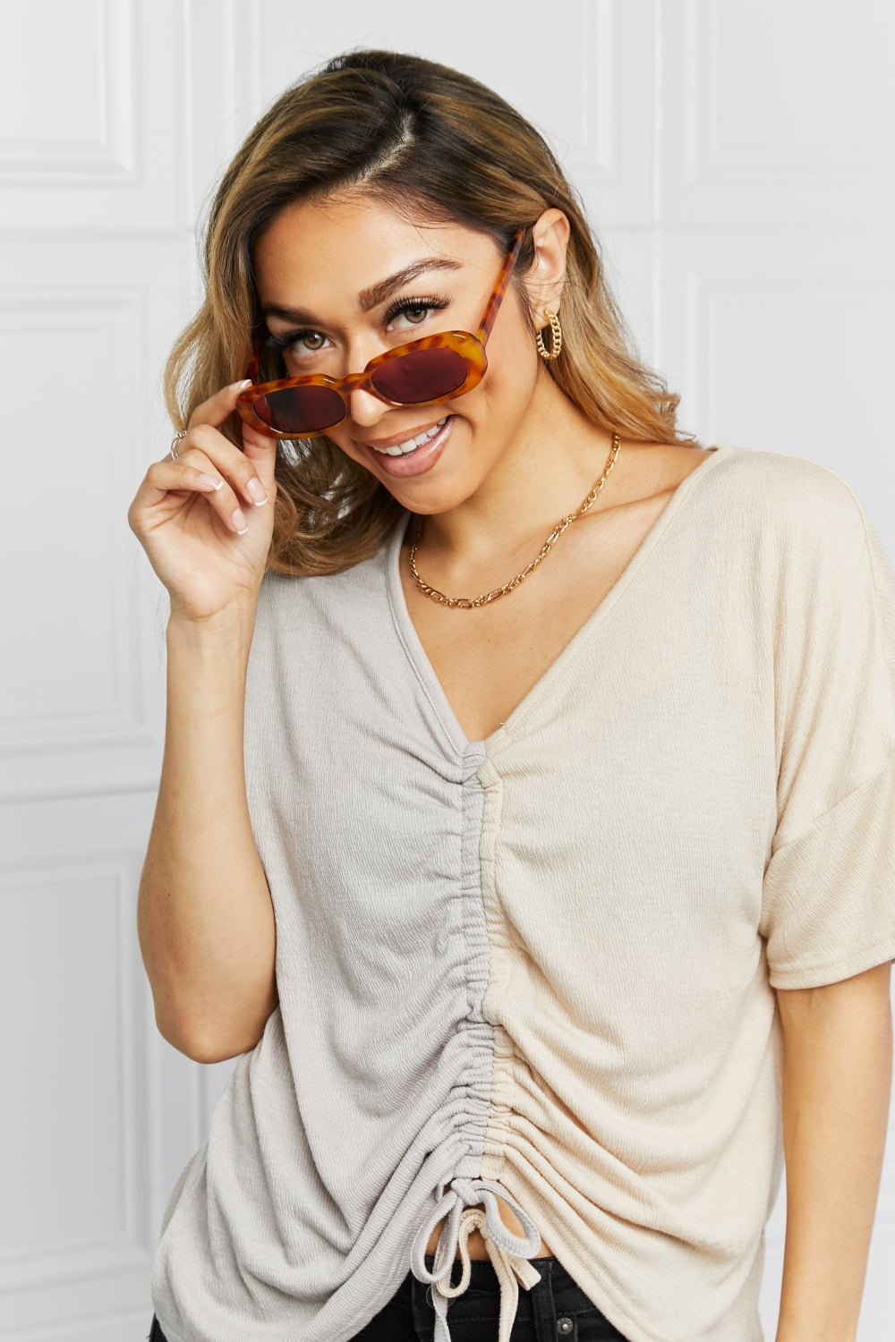 A smiling model adjusts brown tortoiseshell oval full-rim sunglasses, showcasing their softly curved silhouette and dark lenses with a ruched beige top.