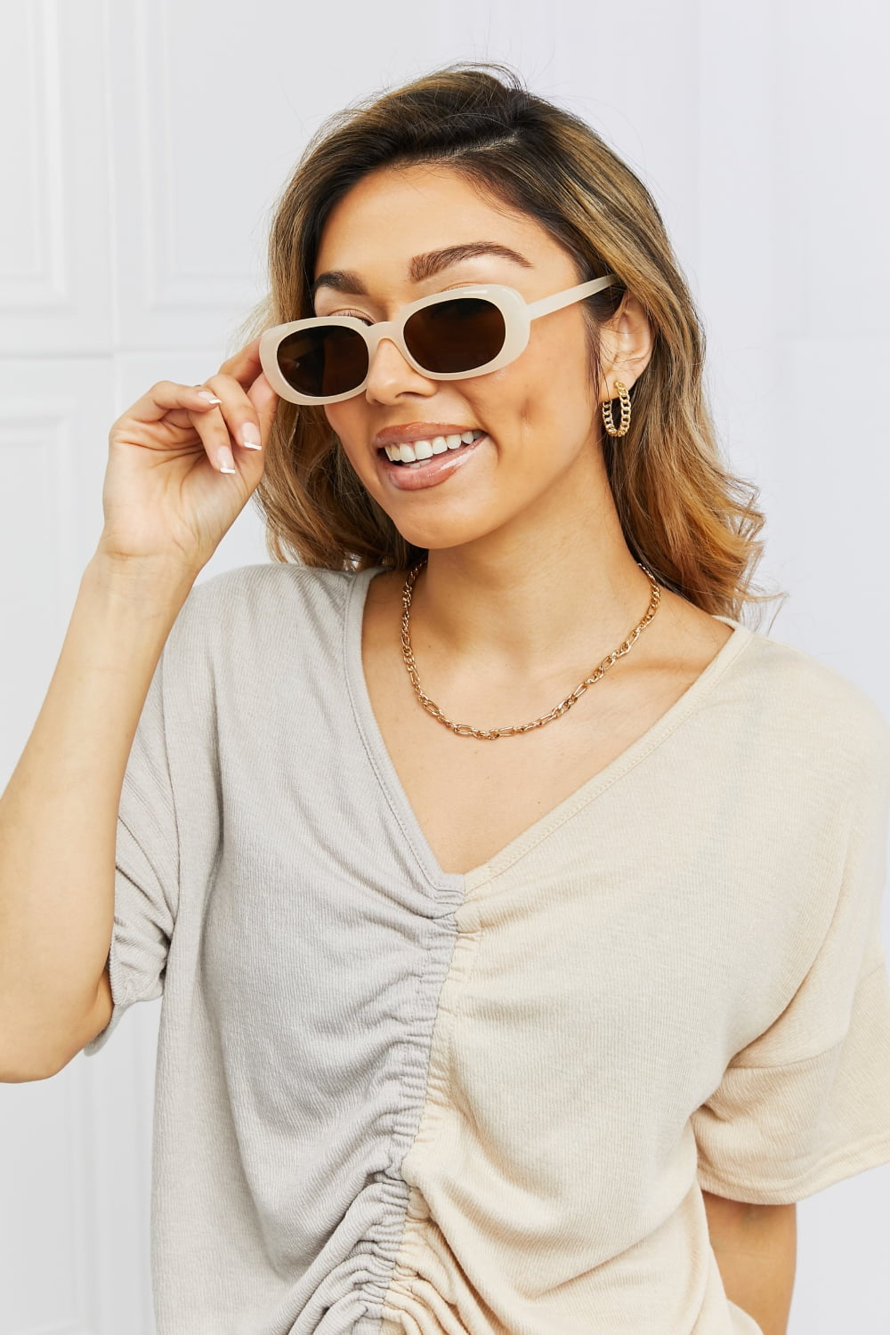 Smiling model adjusts cream oval full-rim sunglasses with dark lenses, showcasing their softly curved silhouette and clean polycarbonate frame.