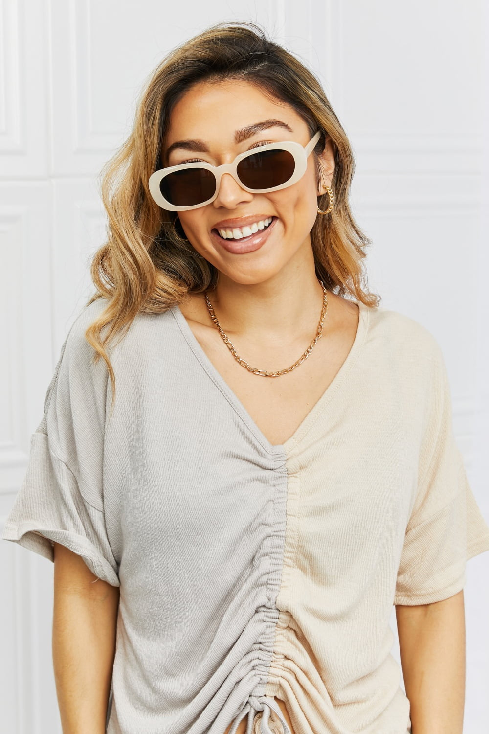 Smiling model faces the camera wearing cream oval full-rim sunglasses with dark brown lenses and a softly curved, vintage-inspired silhouette.