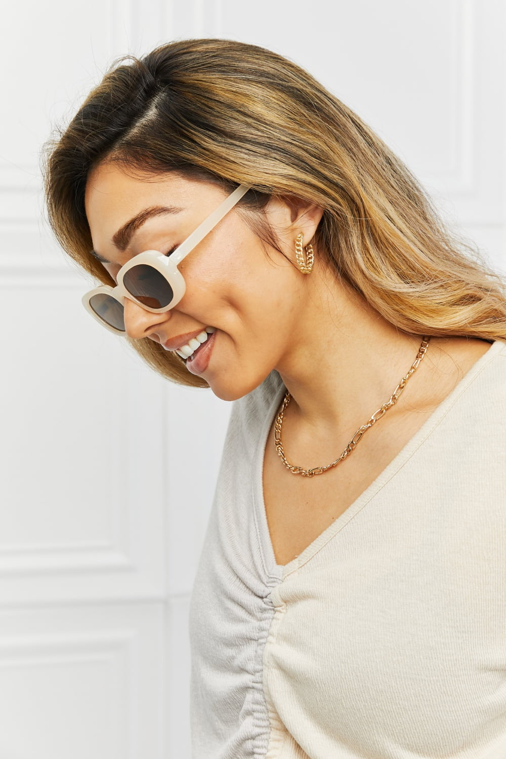 A smiling model tilts her head while wearing cream oval full-rim sunglasses with dark lenses and slim matching temples for a polished vintage-inspired look.
