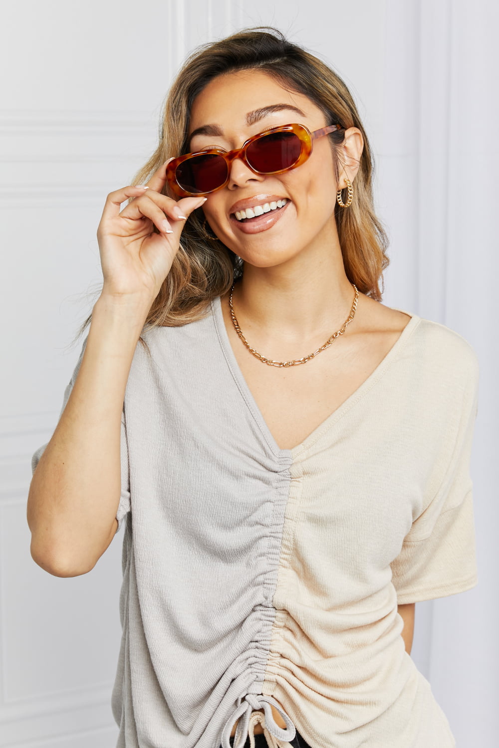A smiling model adjusts tortoiseshell oval full-rim sunglasses while wearing a cream ruched V-neck top, highlighting their vintage-inspired curved silhouette.