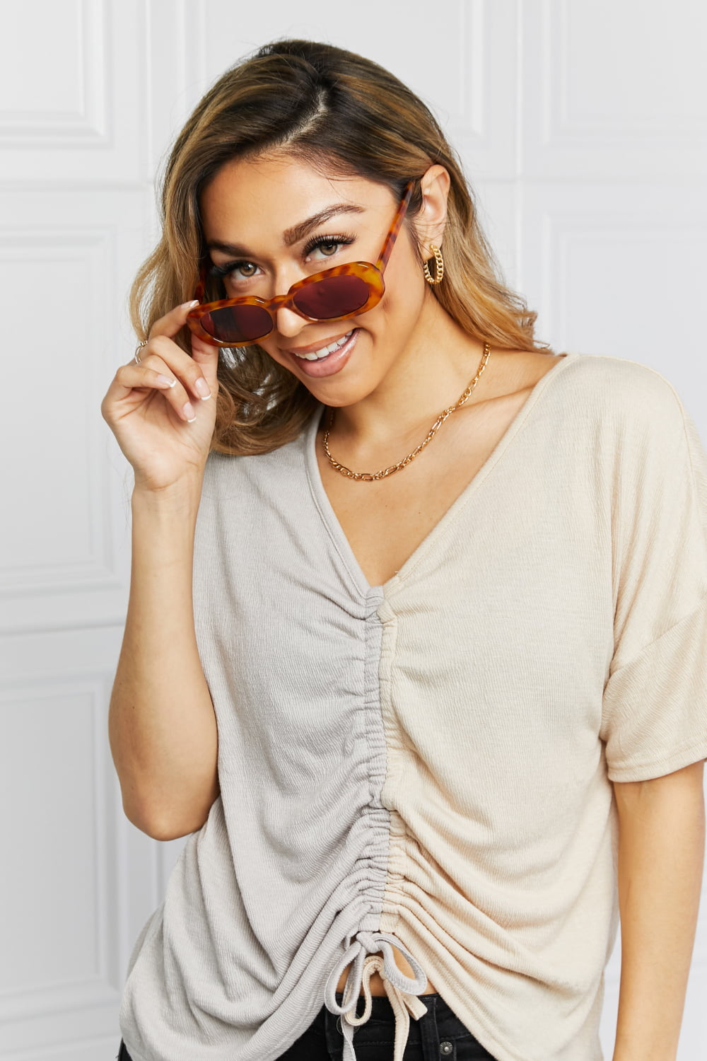 A smiling model adjusts brown tortoiseshell oval full-rim sunglasses, showcasing their softly curved silhouette and dark lenses with a ruched beige top.