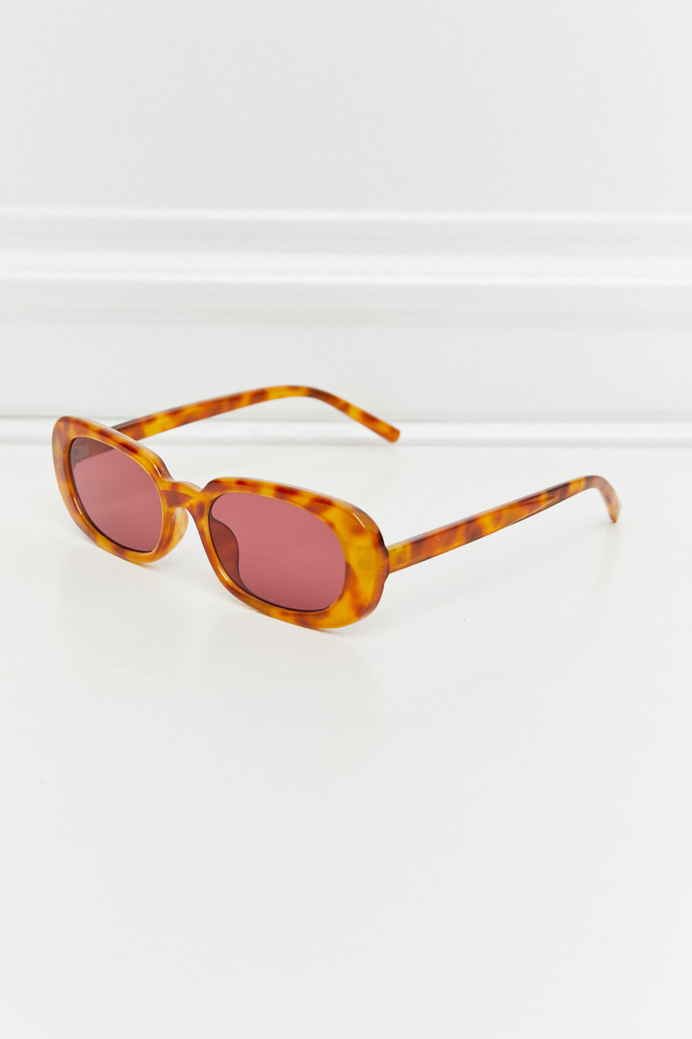 Tortoiseshell oval full-rim sunglasses feature warm amber-brown polycarbonate frames, softly curved pink lenses, and slim temples displayed open at an angle.