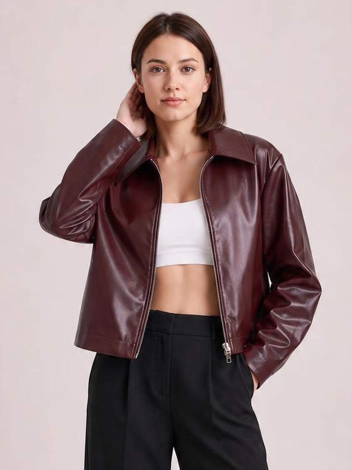 Model wears an oversized burgundy faux leather moto jacket with a zip front and wide collar, posing with one hand at her hair and one in a pocket.