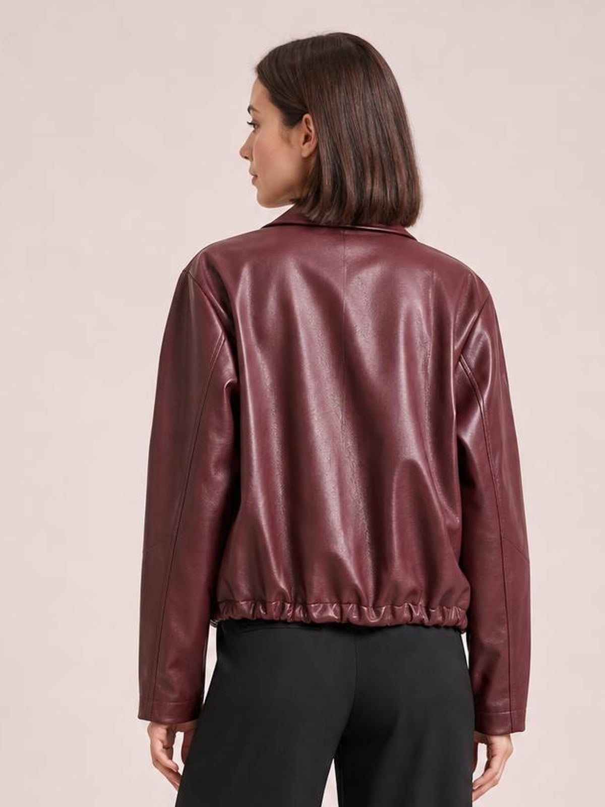 A model shows the back of an oversized burgundy faux leather moto jacket with long sleeves, a pointed collar, seam details, and an elasticized hem.