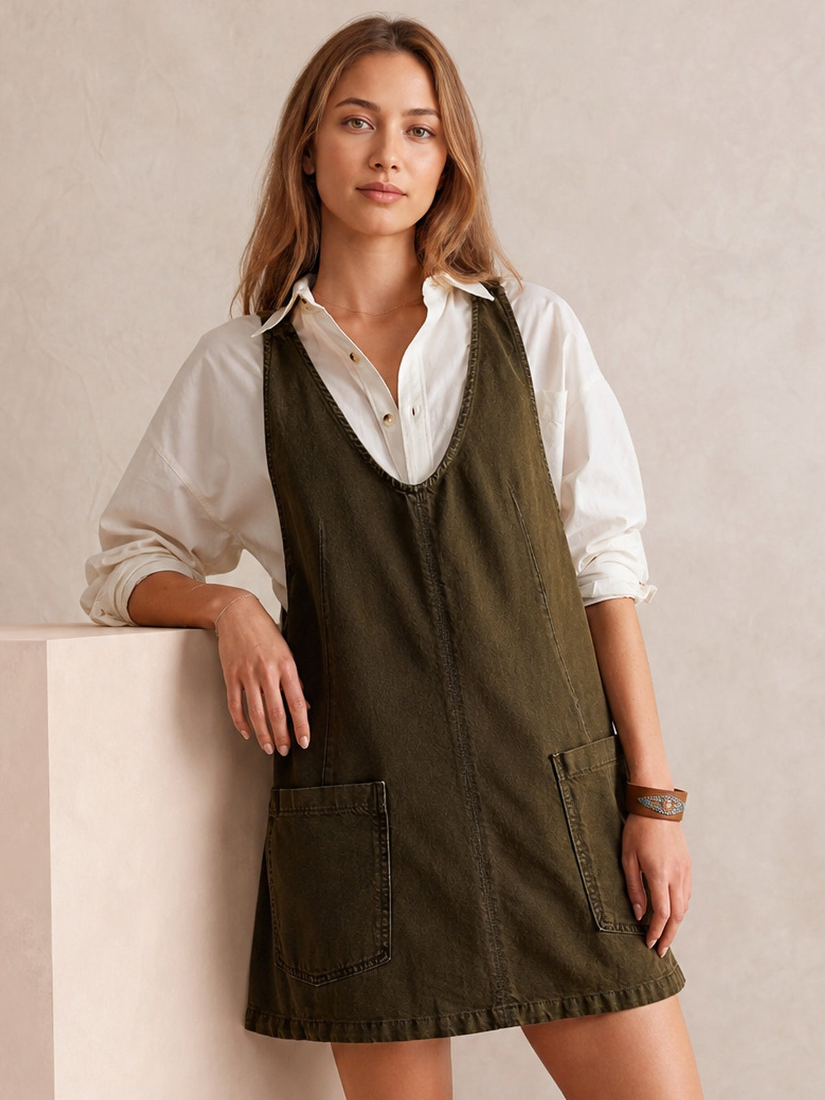 A model leans against a pedestal wearing a dark olive oversized denim tank dress with a deep V-neck, sleeveless cut, A-line silhouette, and front patch pockets.