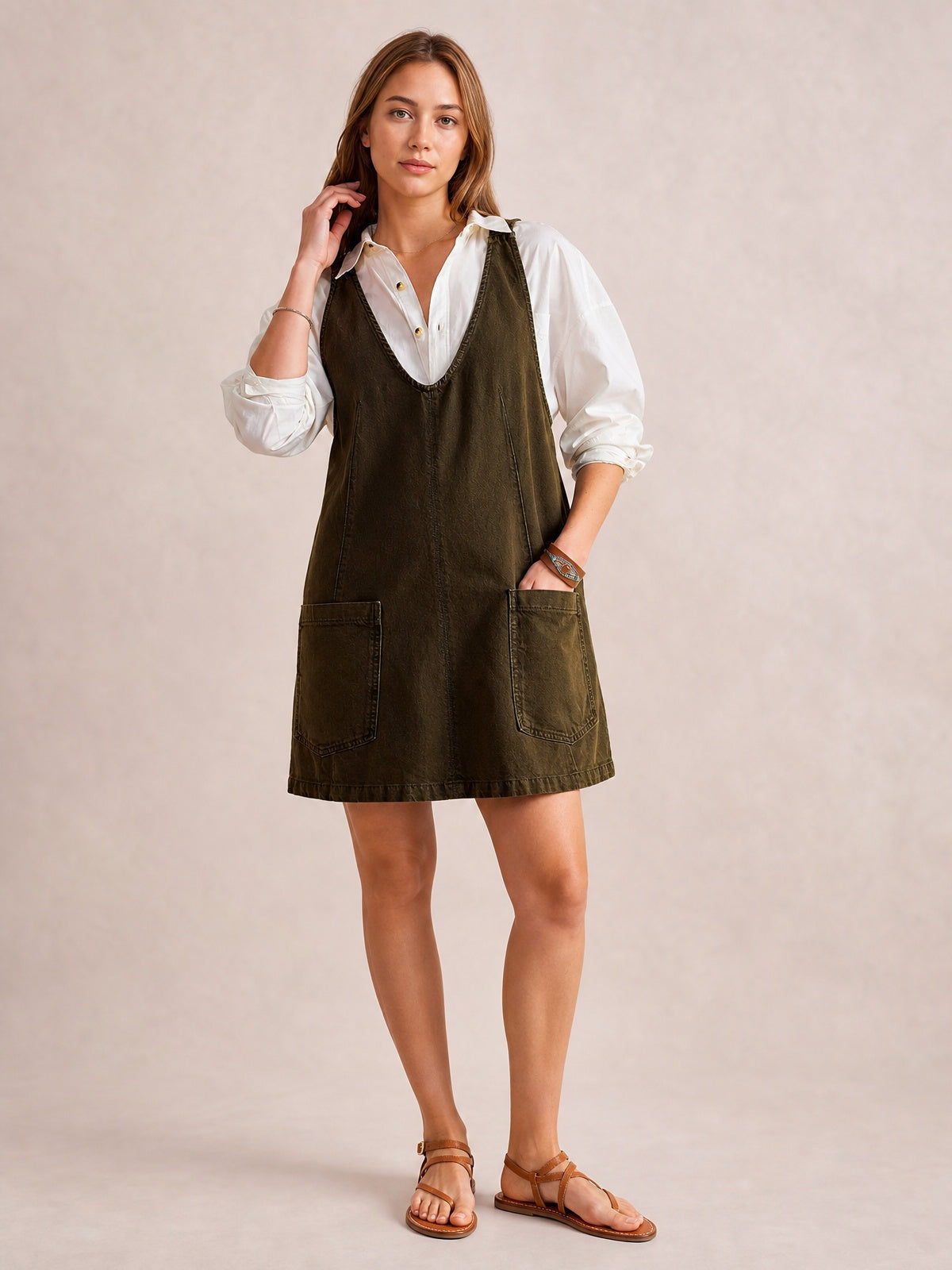 Model stands in an olive denim tank dress with a deep V-neck, oversized A-line silhouette, sleeveless construction, and two front patch pockets over a white shirt.