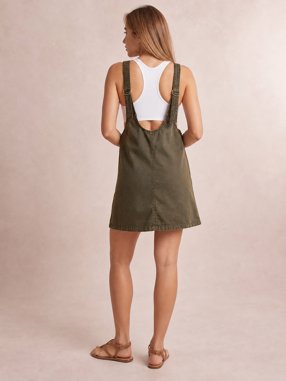 A model stands facing away in an oversized olive denim tank dress with wide adjustable straps, metal buckles, a relaxed A-line silhouette, and an above-the-knee hem.