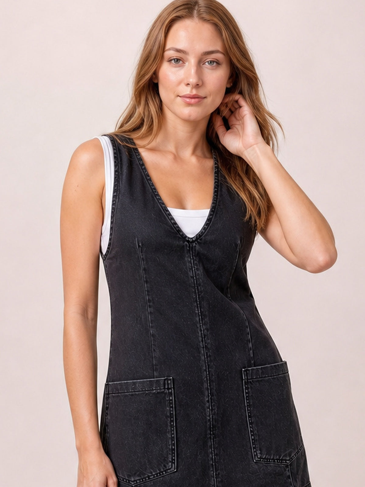 Model poses in a black oversized V-neck denim tank dress with contrast stitching, vertical seaming, and two large front patch pockets over a white top.