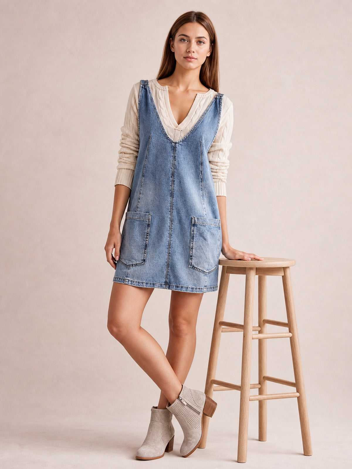 Model poses with one hand on a stool and ankles crossed, wearing a light blue oversized V-neck denim tank dress with front patch pockets and an above-knee silhouette.
