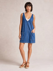 Model poses in an oversized blue denim tank dress with a deep V-neck, sleeveless cut, front patch pockets, structured seams, and an above-the-knee hem.