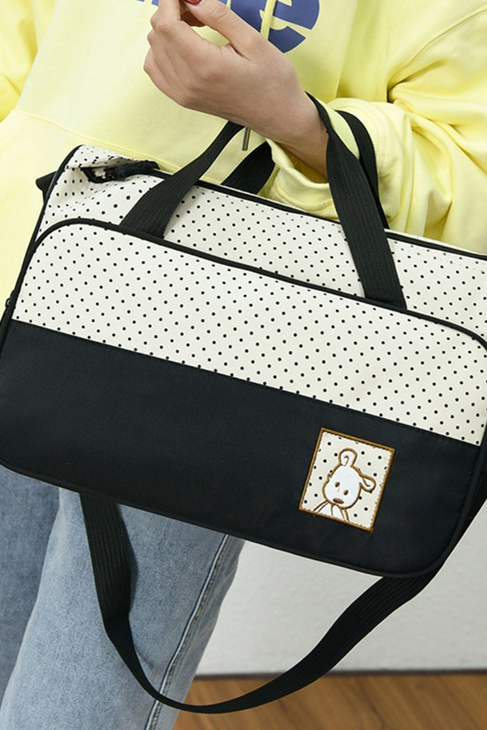 A model holds a softly structured Oxford cloth tote with black contrast panels, polka-dot detailing, dual top handles, and a long shoulder strap.