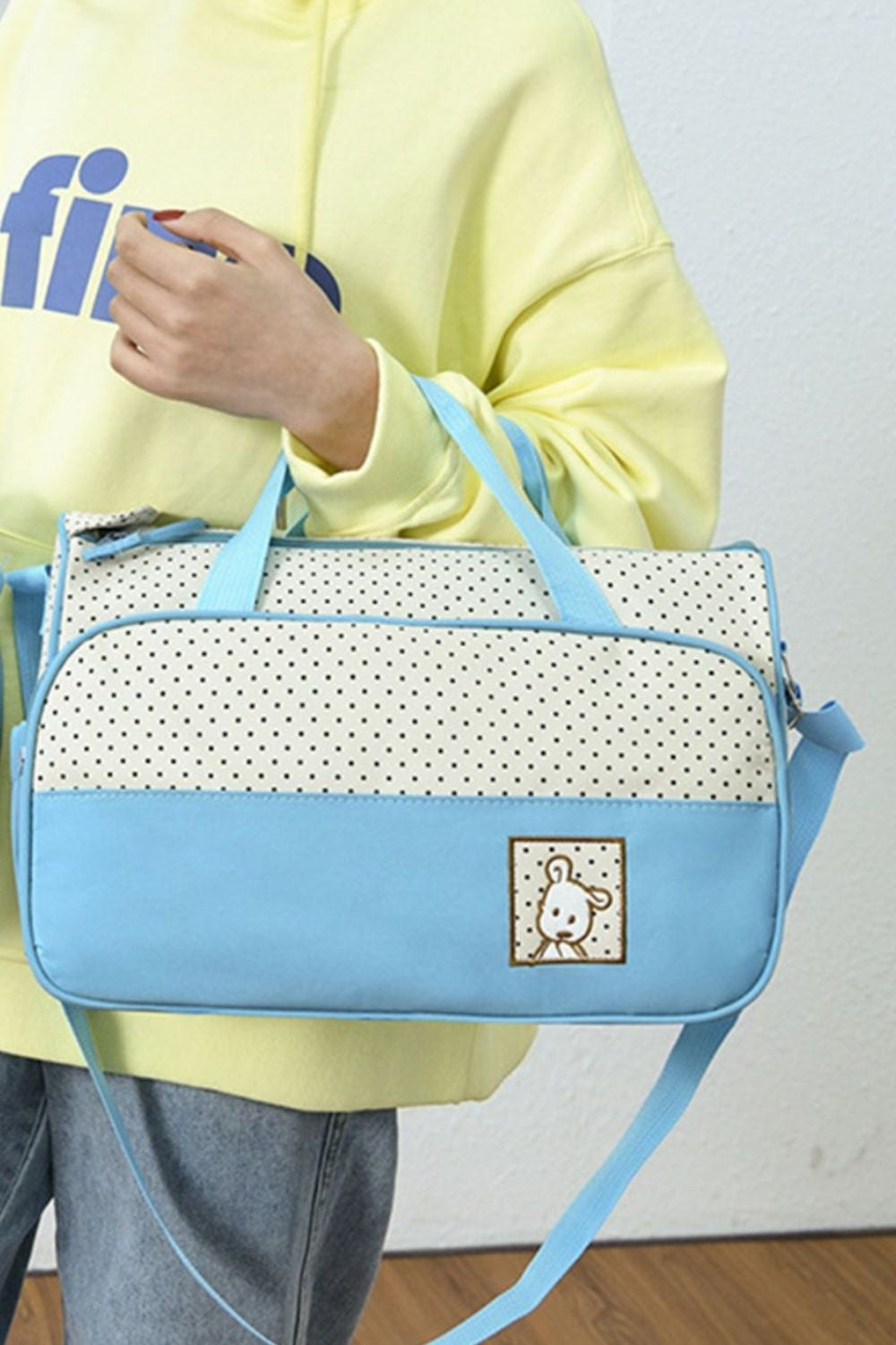 A model holds a softly structured Oxford cloth tote with blue contrast trim, polka-dot panels, two top handles, a long shoulder strap, and a front pocket.
