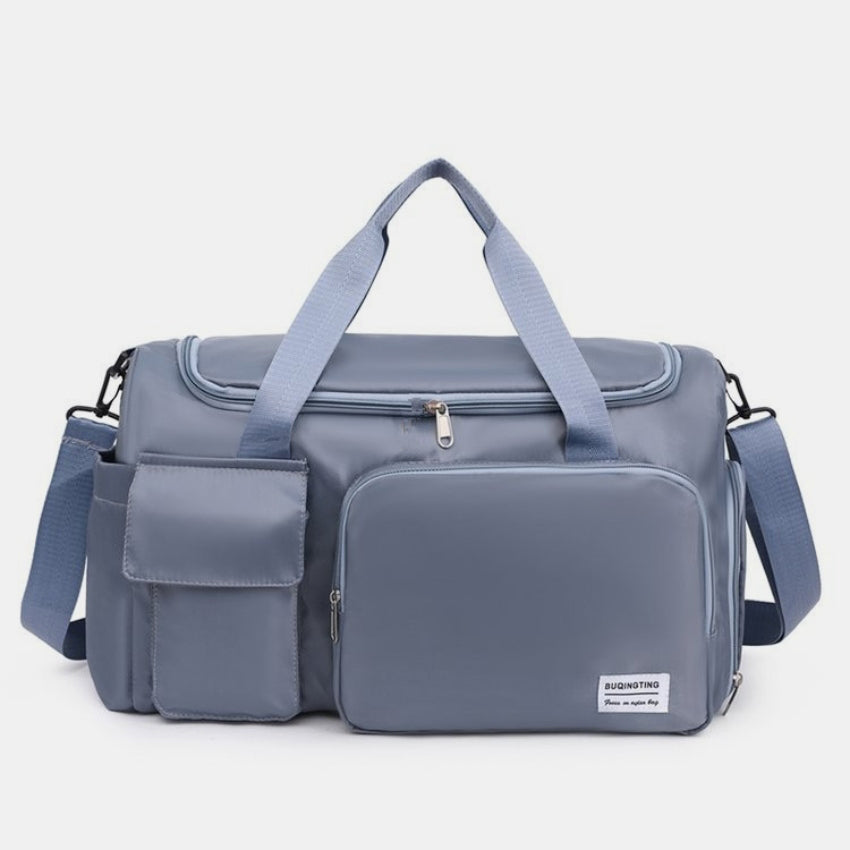 Oversize blue-gray oxford cloth travel bag with a structured silhouette, dual top handles, detachable shoulder strap, and multiple zippered and flap-front pockets.