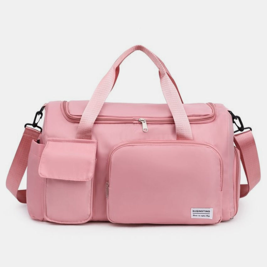 An oversize pink oxford cloth travel bag with a structured silhouette, dual top handles, detachable shoulder strap, wide zip opening, and front zippered and flap pockets.