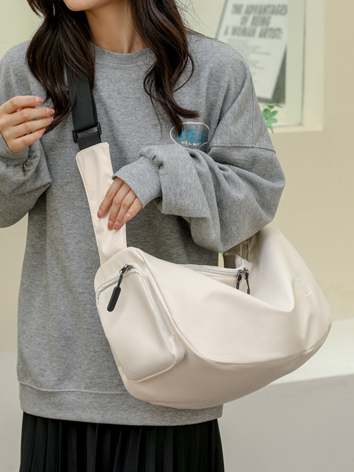 A model in a gray oversized sweatshirt and black pleated skirt holds a large cream Oxford cloth crossbody bag with a slouchy silhouette, wide strap, and zip compartments.