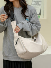 A model in a gray oversized sweatshirt and black pleated skirt holds a large cream Oxford cloth crossbody bag with a slouchy silhouette, wide strap, and zip compartments.