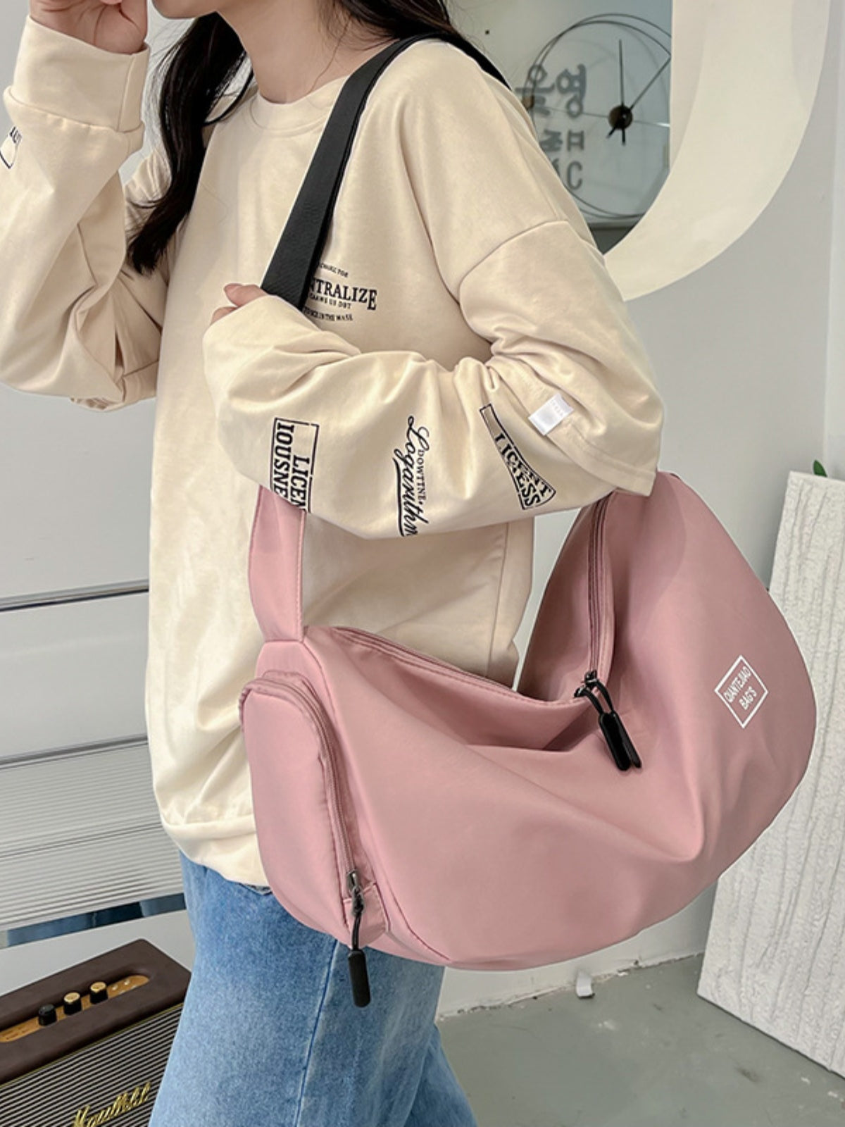 A model in a cream sweatshirt and jeans wears a large pink Oxford cloth crossbody bag with a slouchy curved silhouette, long strap, and zip compartments.