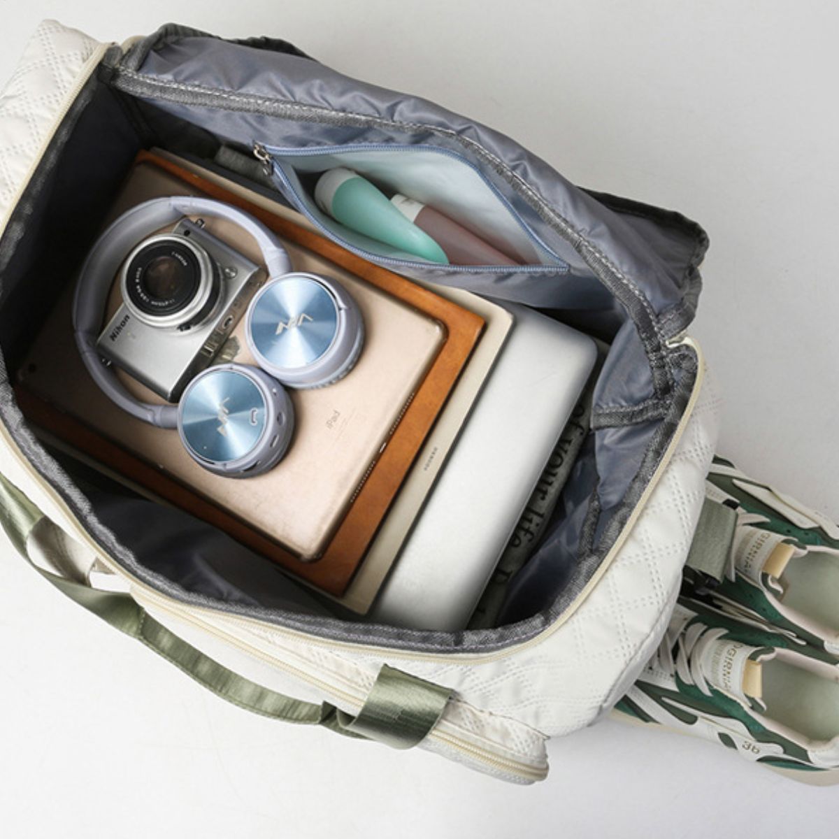 An oversize white Oxford cloth travel bag sits open with green handles, a roomy zip-top interior, and a visible zip pocket holding organized essentials.