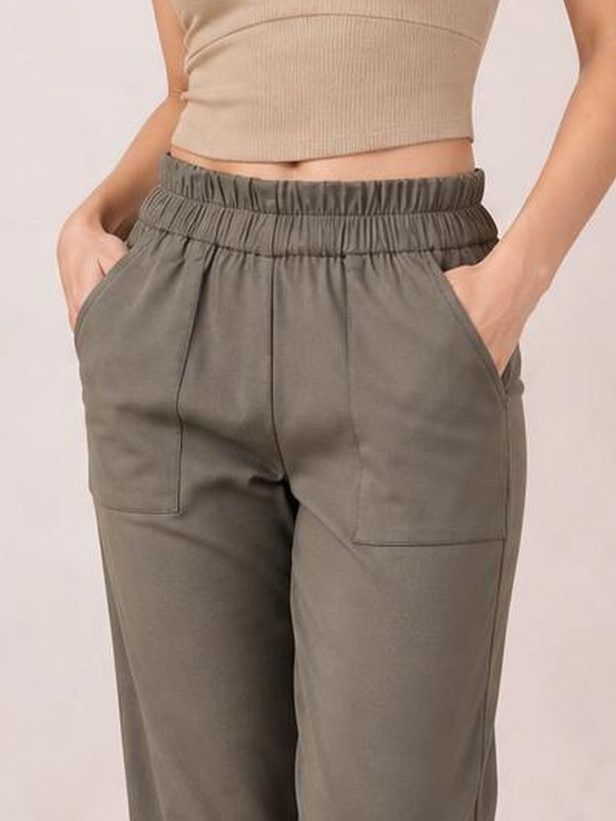 Model wearing taupe paperbag waist pants with an elastic gathered waistband, visible side pockets, and a relaxed straight-leg silhouette.