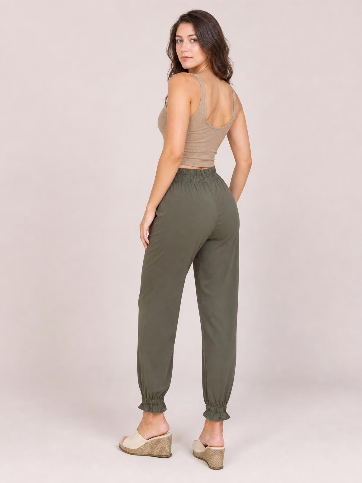 A model turns to show olive-green paperbag waist pants with side pockets, a relaxed tapered silhouette, gathered waistband, and elastic ankle cuffs.
