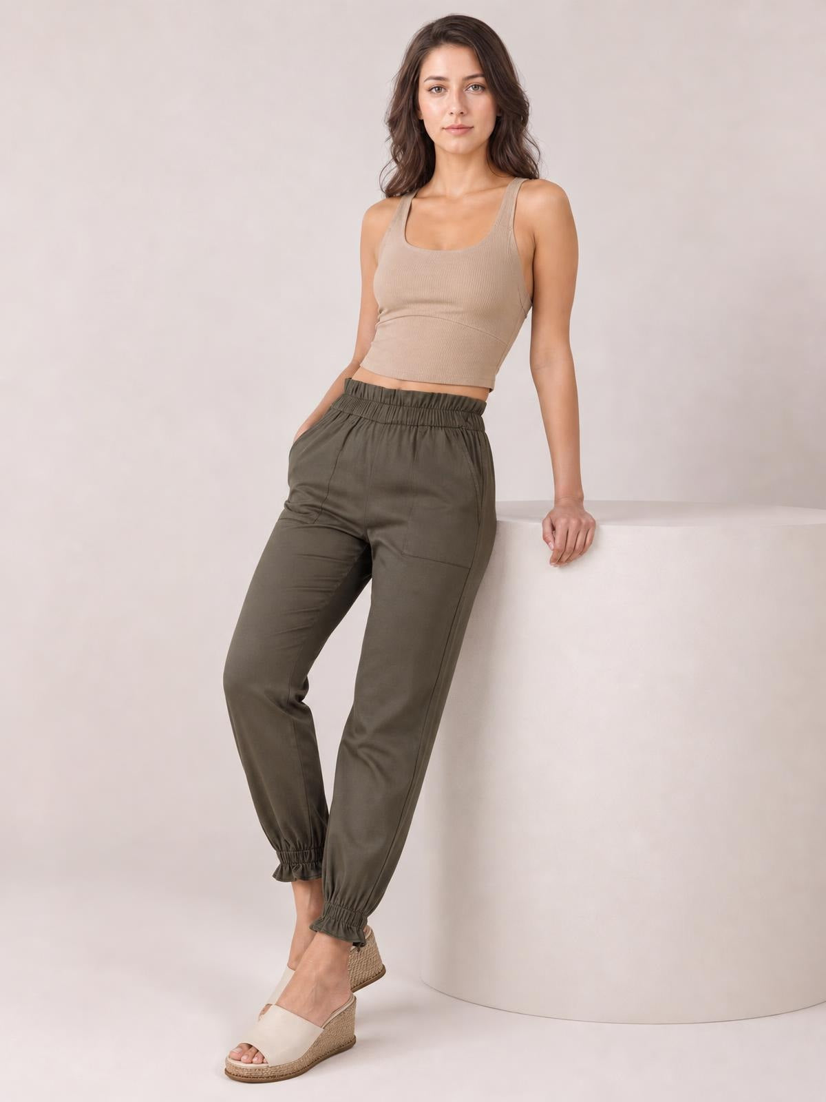 A model leans against a ledge wearing olive paperbag waist pants with pockets, a gathered high waist, tapered silhouette, and elastic ankle cuffs.