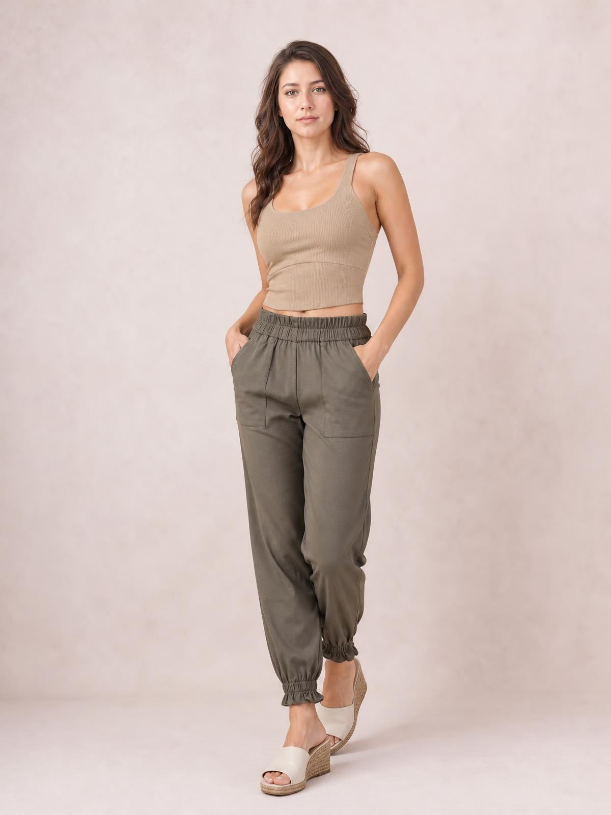Model poses with hands in the pockets of olive paperbag waist pants featuring a gathered elastic waistband, relaxed tapered legs, and elastic cuffs.
