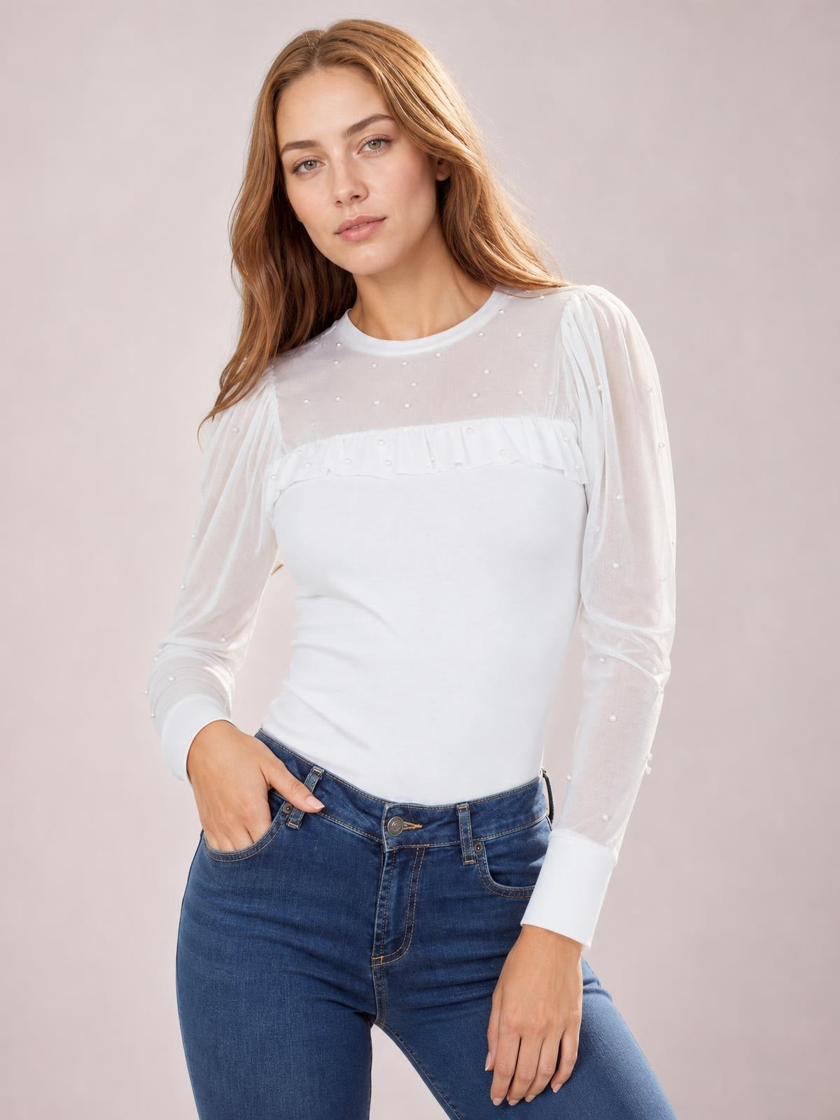 A model poses with one hand in her jeans pocket, wearing a white pearl-decorated top with sheer mesh long sleeves, a ruffled yoke, and fitted ribbed silhouette.