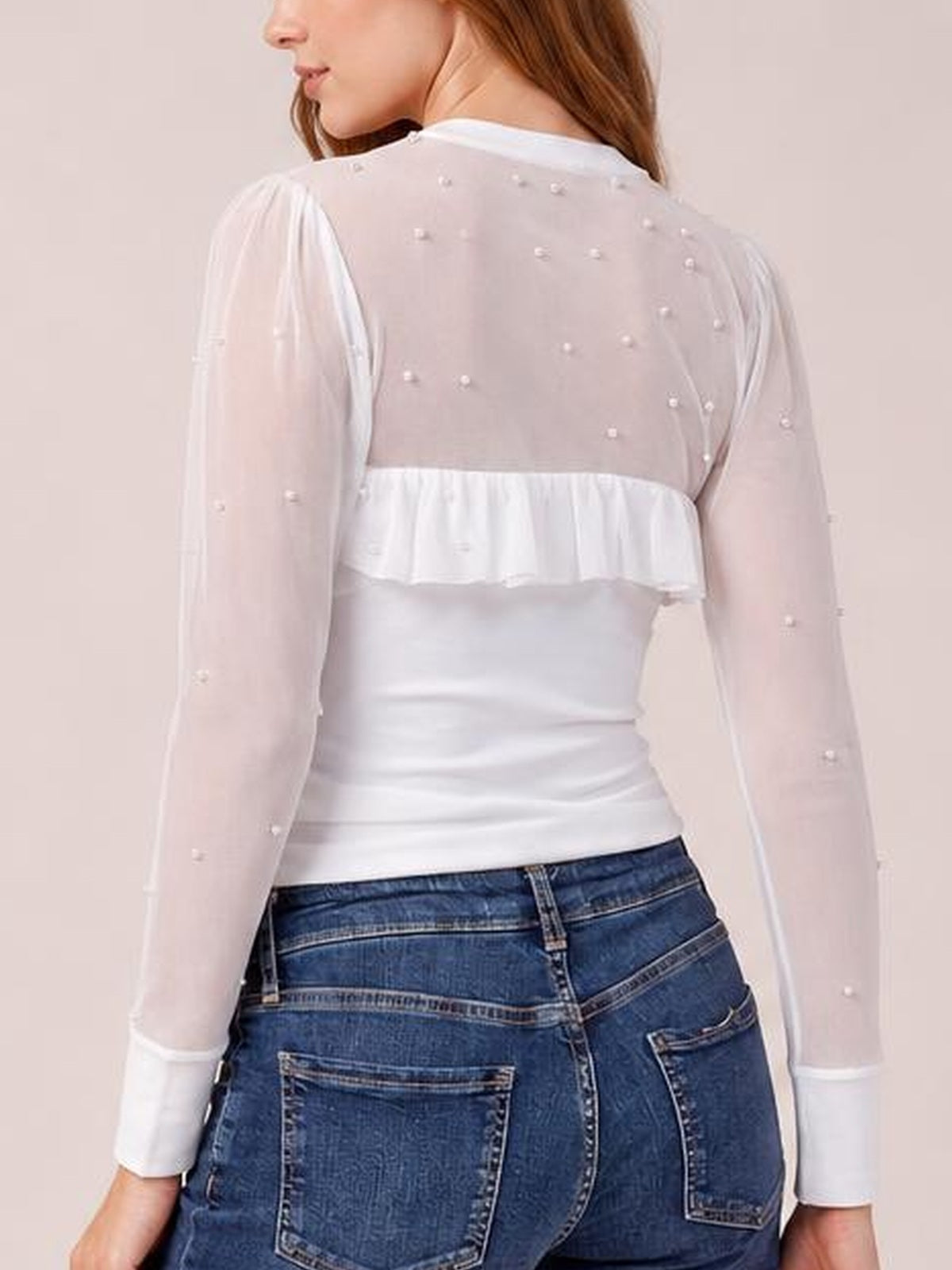 Back view of a model wearing a white pearl-decorated semi-sheer mesh top with long sleeves, a solid fitted bodice, and a ruffled waist panel.