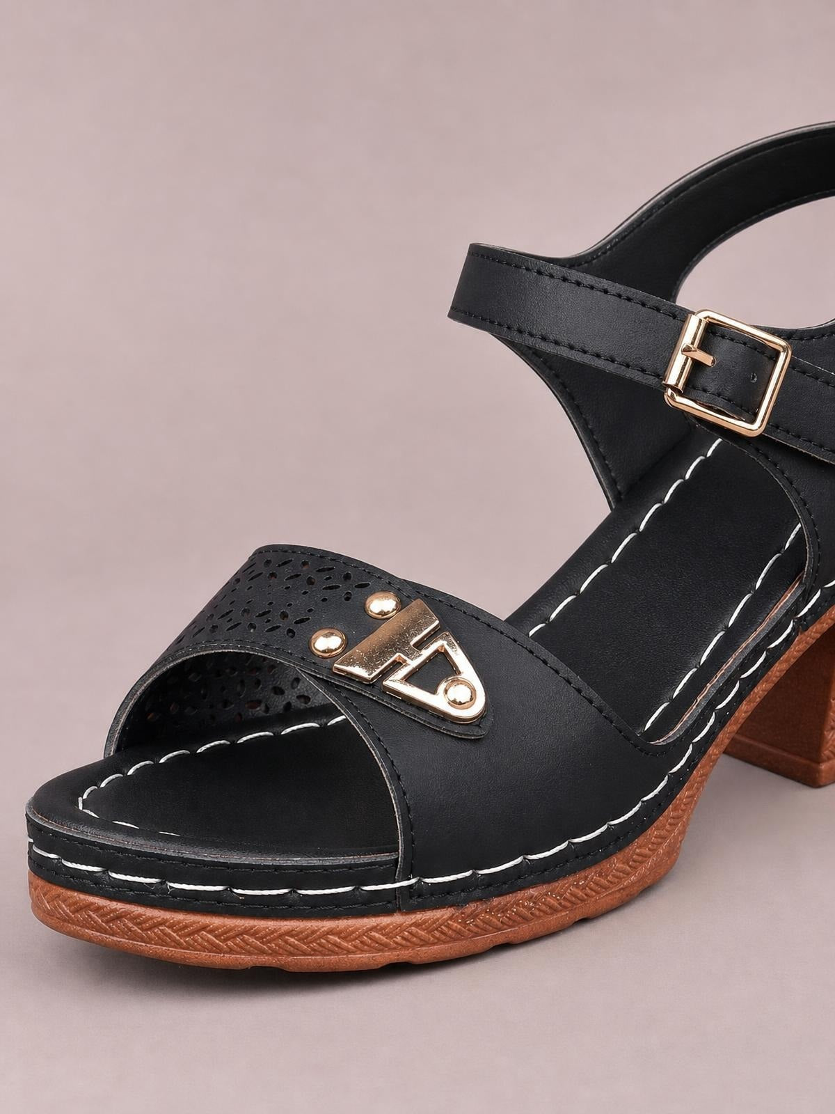 Black peep toe block heel sandals feature a wide perforated strap, gold-tone embellishment, ankle buckle, white stitching, and a brown mid-height sole.