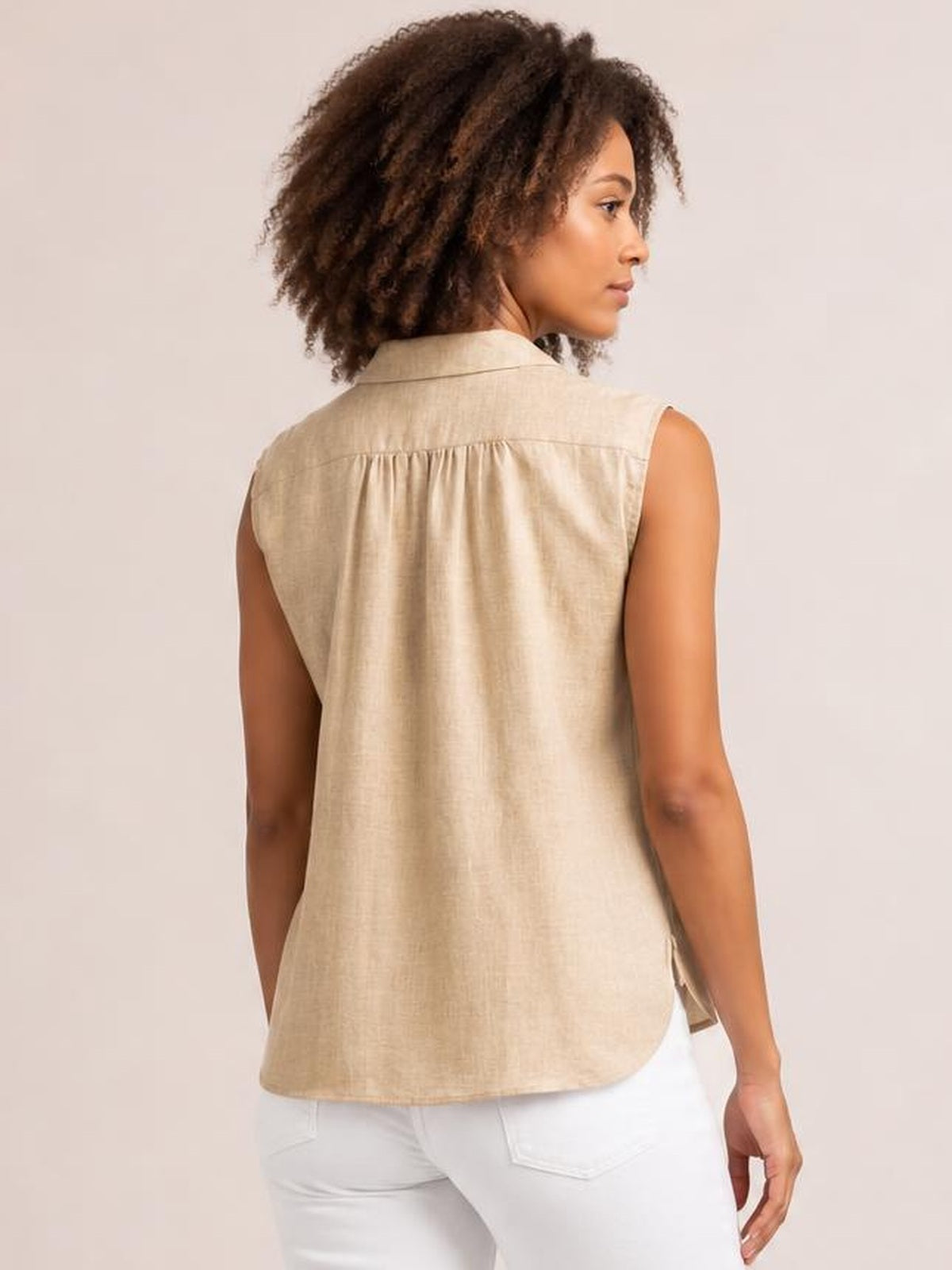 Model shown from behind in a relaxed pose wearing a beige button-up collared neck cap sleeve shirt with a loose silhouette and gathered back yoke.