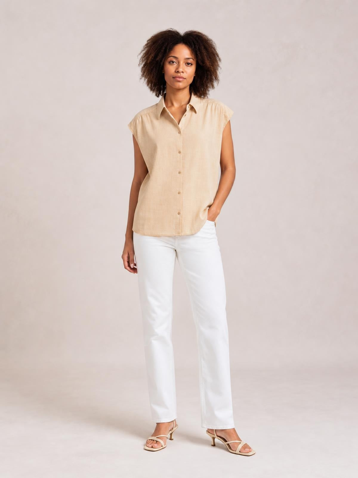 Model poses in a relaxed stance wearing a beige button-up collared neck cap sleeve shirt with a straight silhouette and rounded hem.