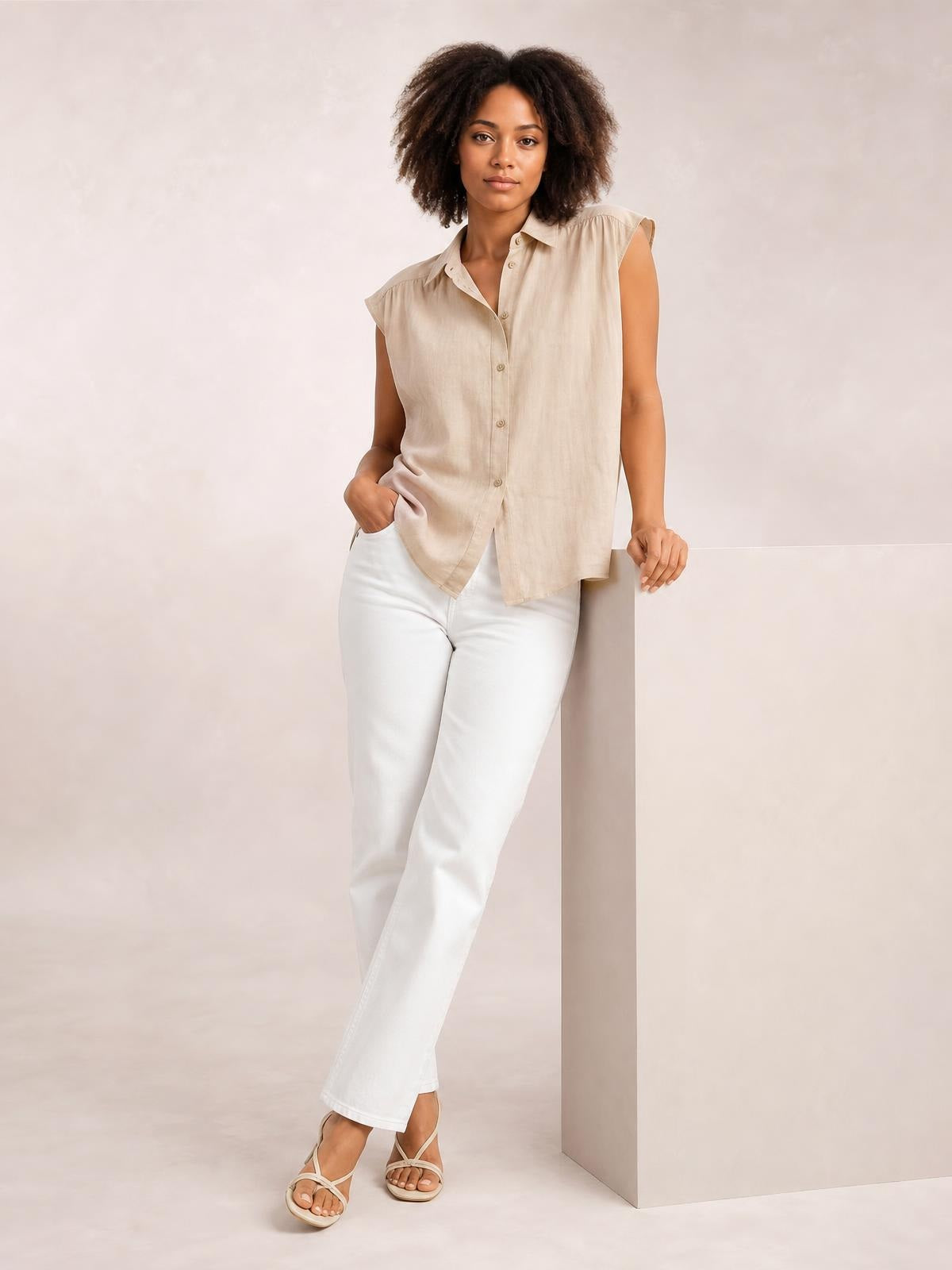 Model poses in a beige button-up collared neck shirt with cap sleeves, a relaxed straight silhouette, curved hem, and visible front buttons.