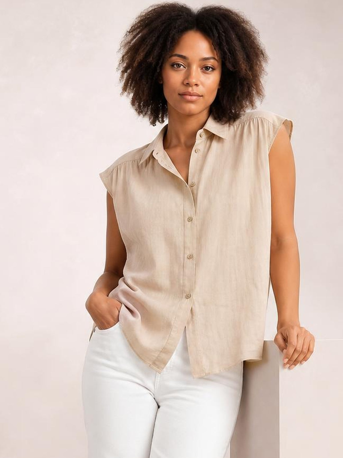 Model wears a beige button-up shirt with a collared neck, cap sleeves, relaxed silhouette, and curved hem, posing with one hand in her pocket.
