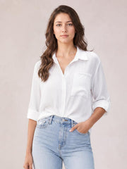 A model stands in a white collared neck twisted shirt with a relaxed silhouette, button front, chest pocket, and rolled three-quarter sleeves.