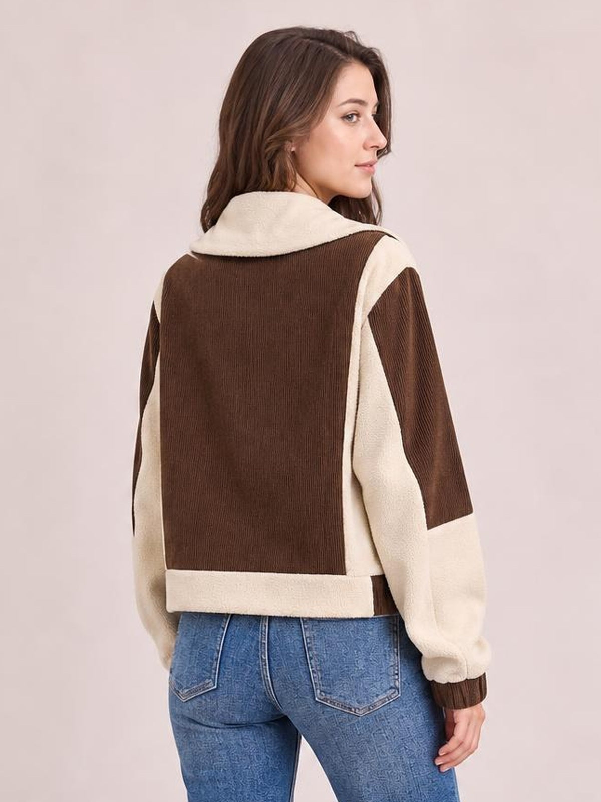 A model looks over her shoulder wearing a brown-and-cream color block jacket with a hood, contrasting sleeves, ribbed cuffs, and a cropped relaxed silhouette.
