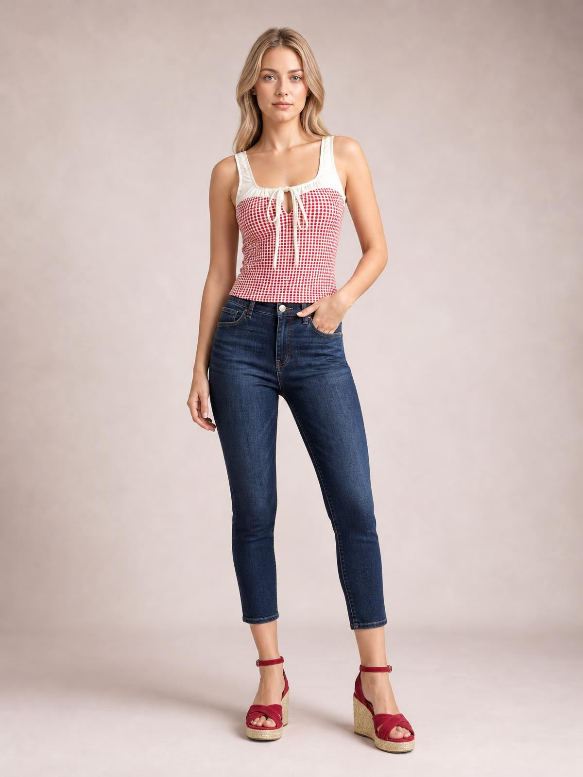 A model stands with one hand in her jeans pocket, wearing a fitted red-and-white contrast plaid square neck tank with white straps and a tied front.