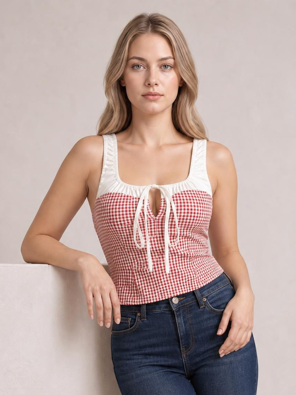 A model leans against a ledge wearing a fitted red-and-white contrast plaid tank with a square neck, wide straps, and a front tie detail.