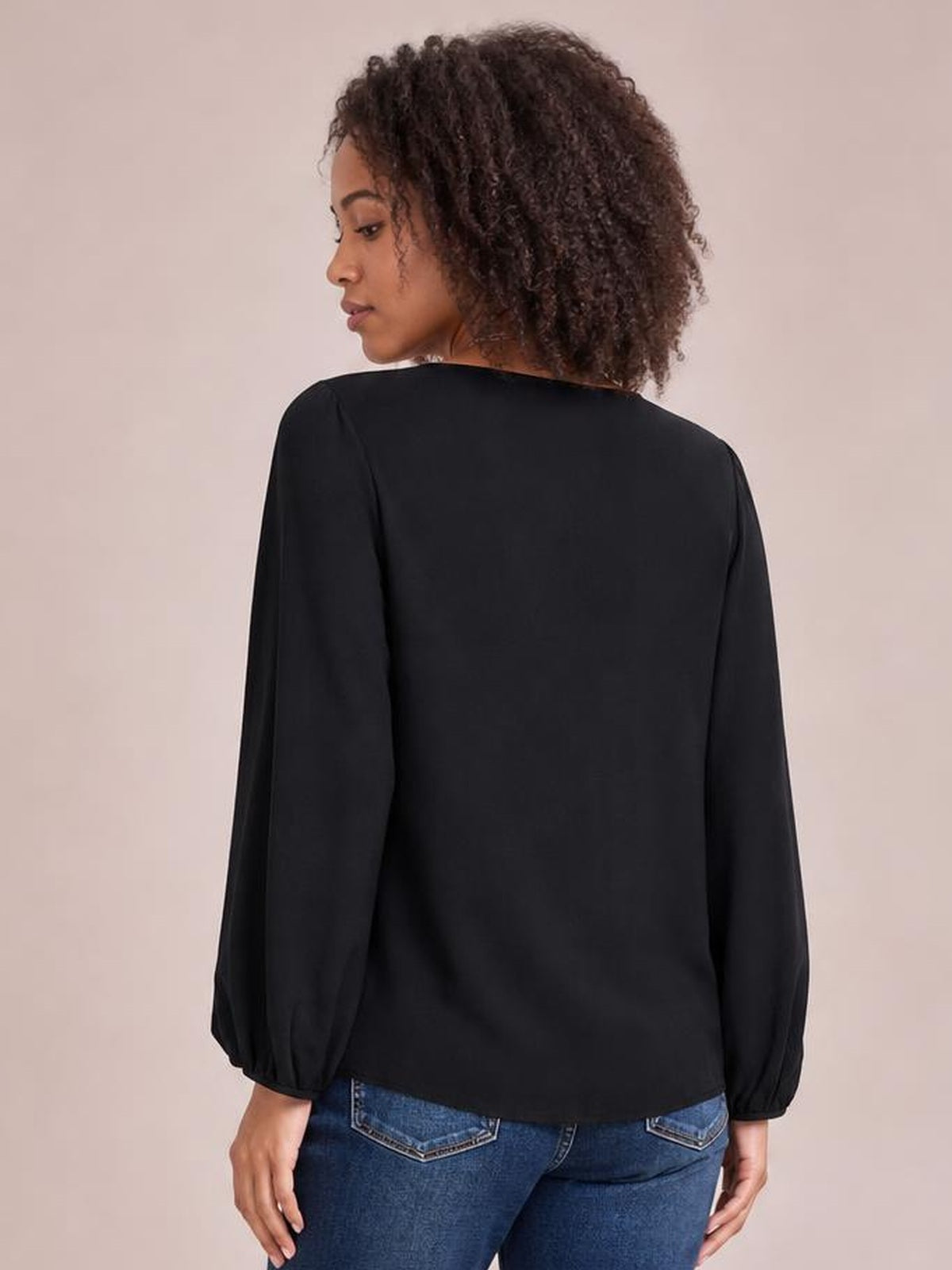 Model shown from behind wearing a black long-sleeve blouse with a relaxed straight silhouette and softly gathered cuffs.