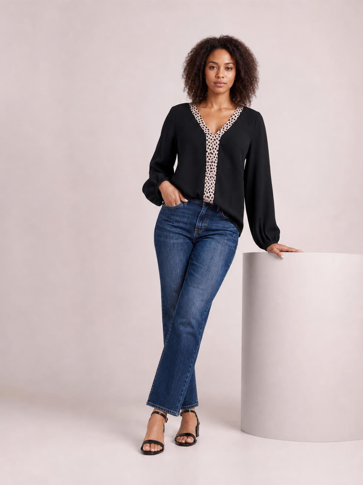 A model wears a black contrast V-neck long sleeve blouse with animal-print neckline and front trim, a relaxed silhouette, and gathered cuffs, posing with one hand in her jeans pocket.