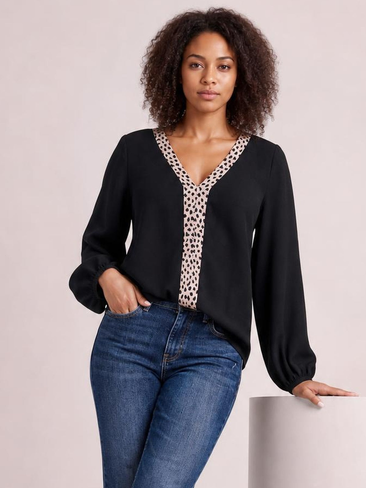 Model wearing an opaque black contrast V-neck long sleeve blouse with a patterned trim and relaxed silhouette, posing with one hand in her jeans pocket.